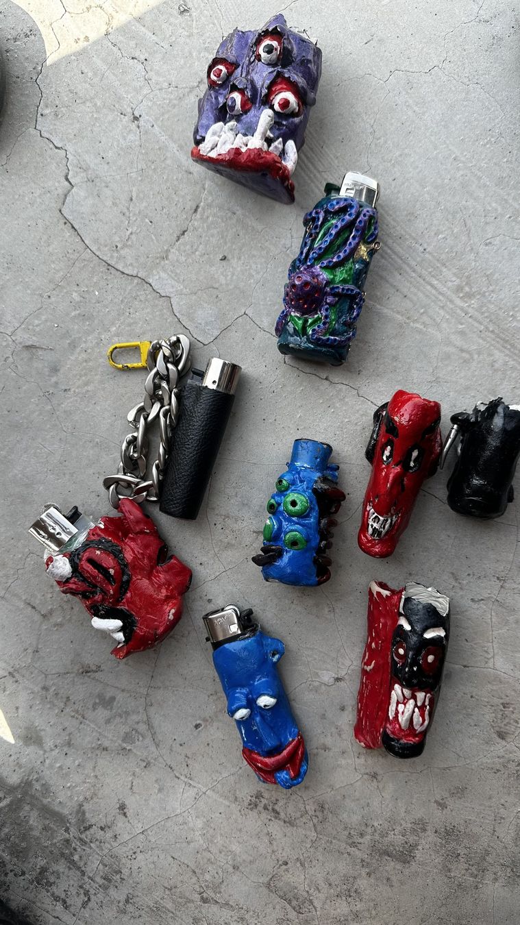 The MUDDEALER gang: nine handmade clay lighter cases laid out on a concrete floor, with purple, red, blue and black monster faces.
